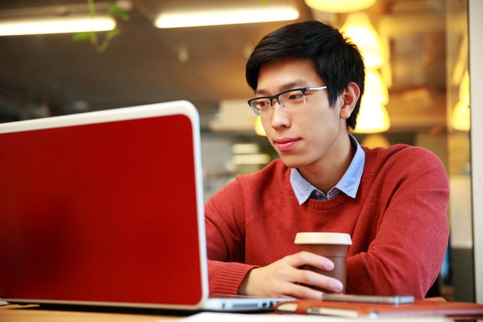 Young asian man in glasses working on laptop and holding cup of coffee